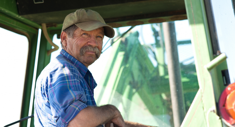 一位年长的农民坐在一辆绿色拖拉机的驾驶室里，回头看着镜头. 他留着浓密的灰胡子，戴着一顶棕褐色的帽子，穿着一件蓝色格子衬衫.